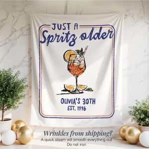 May include: White fabric banner featuring a cocktail illustration and the text "JUST A Spritz older OLIVIA'S 30TH EST. 1996". The banner has a blue and red border and a drawing of an orange cocktail.