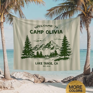 May include: A beige and white striped tapestry with the text "WELCOME TO CAMP OLIVIA" and "LAKE TAHOE, CA EST. 2026". The tapestry features a green illustration of mountains, trees, and clouds.