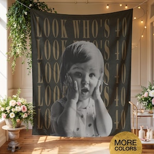 May include: A dark grey rectangular wall tapestry featuring the repeating gold text LOOK WHO'S 40 and a large black and white photograph of a young child with hands pressed against their cheeks in a surprised expression, centred on the fabric.