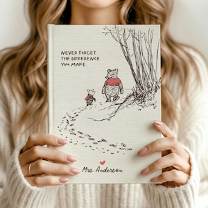 May include: A cream-coloured hardback book featuring a black ink illustration of Winnie the Pooh and Piglet walking near trees. The text reads, "NEVER FORGET THE DIFFERENCE YOU MAKE." and "Mrs. Anderson" with a small red heart icon below.
