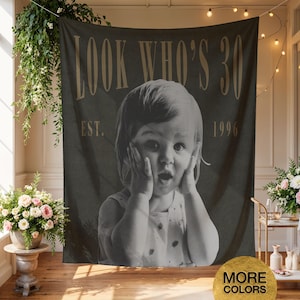 May include: A dark gray rectangular wall tapestry featuring a black and white photograph of a child with hands on their cheeks. The text reads LOOK WHO'S 30 at the top and EST. 1996 at the sides in a gold serif font. The fabric hangs vertically.