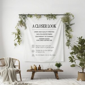 May include: A white polyester fabric wall hanging with the text "A Closer Look" and care instructions. The fabric is displayed on a black rod, with greenery and a wooden bench with decorative items in front of it.