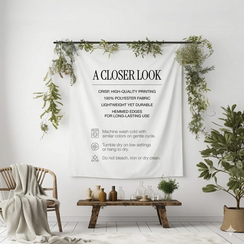 May include: A white polyester fabric wall hanging with the text "A Closer Look" and care instructions. The fabric is displayed on a black rod, with greenery and a wooden bench with decorative items in front of it.
