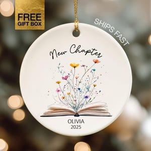 May include: A white ceramic ornament with a gold hanging string. The ornament features the words "New Chapter" and a watercolor illustration of flowers emerging from an open book. The name "Olivia" and the year "2025" are printed below the book.