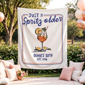 May include: A white banner with a blue and red border, displaying the text "JUST A Spritz older" and "OLIVIA'S 30TH EST. 1996". The banner features a cocktail graphic with an orange slice and a straw. Pillows and balloons are in the background.