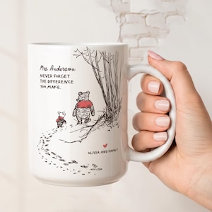 May include: A white ceramic mug featuring a black ink illustration of Winnie the Pooh and Piglet walking along a path. The text reads Mrs. Anderson NEVER FORGET THE DIFFERENCE YOU MAKE and OLIVIA AND FAMILY with a small red heart icon.