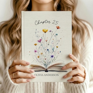 May include: A hardcover book with a cream-colored fabric cover featuring a watercolor illustration of an open book with colorful wildflowers growing from the pages. The text Chapter 25 is printed at the top and Olivia Anderson at the bottom.