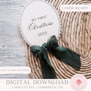 Puede incluir: Adorno navideño ovalado de madera con bordes festoneados, con la inscripción "MY FIRST Christmas 2025". Una cinta verde oscuro está atada debajo del adorno. También se ven las palabras "LASER READY" y "FONT NAMES INCLUDED".