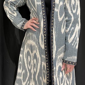 Könnte beinhalten: Eine lange, grau-weiße gemusterte Jacke mit Reißverschluss und dekorativem Besatz. Die Jacke hat eine lockere Passform und einen leicht ausgestellten Saum.