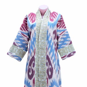 Könnte beinhalten: Eine lange, vorne offene Robe mit einem farbenfrohen Ikat-Muster in Blautönen, Rosa und Weiß. Die Robe hat einen goldenen Stickrand an den Kanten und vorne.