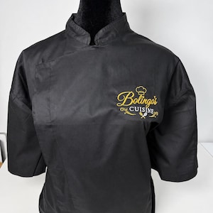May include: A black chef coat with a mandarin collar and elbow-length sleeves. The left chest features yellow embroidered text that reads Bolingo's CUISINE, accompanied by a small chef hat icon and a crossed spoon and spatula graphic.