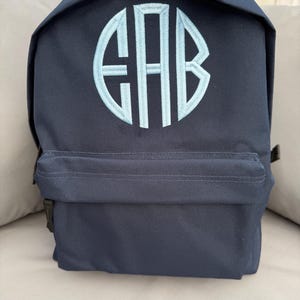 May include: A navy blue backpack with a light blue embroidered monogram. The bag has a front pocket and adjustable straps. The monogram is in a circular design, offering a personalised touch. Suitable for school or travel, this backpack is practical and stylish.