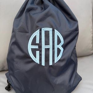 May include: Dark navy drawstring bag with a light blue embroidered monogram. The bag has a rounded shape and a black drawstring closure. The monogram features the letters "EHB" in a stylized font, adding a personalized touch to the bag.