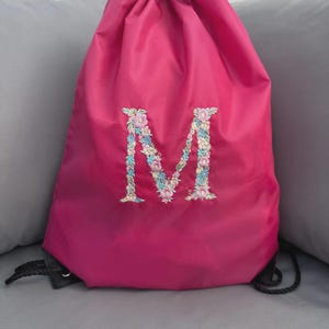 May include: A bright pink drawstring bag with a black drawstring. The letter "M" is embroidered on the front with a floral design in pink, blue, and white. The bag is made of a smooth material, possibly nylon. Ideal for carrying items.