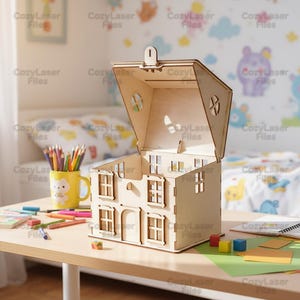 May include: A wooden dollhouse with an open roof and hinged lid is on a table. It has cut-out windows and a door. The scene includes a yellow mug with pencils, coloured blocks, and art supplies, suggesting a craft or play theme.