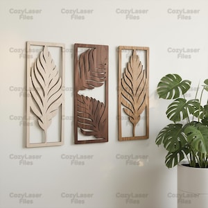 Puede incluir: Tres obras de arte murales de madera con diseños de hojas. Las piezas son rectangulares con un recorte de hoja en el centro. Los tonos de madera varían de claro a marrón oscuro. El arte está montado en una pared blanca, con una planta en maceta en el fondo.