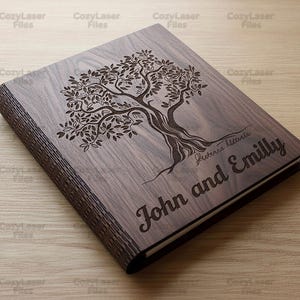 May include: A wooden book with a tree design engraved on the cover. The names "John and Emilly" are engraved below the tree. The book is made of dark wood and has a natural wood grain.