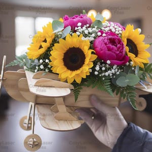 May include: A wooden airplane-shaped flower holder filled with sunflowers, pink peonies, and white baby's breath. The wooden plane has a propeller and wheels. The flowers are vibrant and fresh, creating a cheerful display.