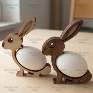 May include: Two wooden Easter bunny egg holders. One is light-colored, the other is dark brown. Each bunny holds a white egg. The bunnies are made of laser-cut wood and are on a light-colored surface.
