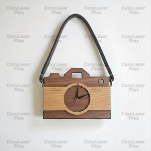 May include: A wooden camera-shaped wall clock with a dark brown body and a lighter wood panel. The clock face is circular with black hands. A grey strap is attached to the top. The text "CozyLaser Files" is repeated on the white background.