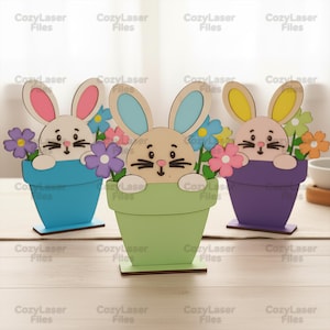 May include: Three wooden Easter bunny decorations. Each bunny is in a different coloured flower pot: blue, green, and purple. The bunnies have different coloured ears and are surrounded by colourful flowers. The text "Cozy Laser Files" is repeated throughout the image.