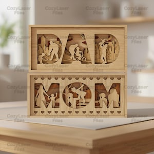 May include: Two wooden laser-cut signs, one reading "DAD" and the other "MOM." The "DAD" sign features silhouettes of a father and child playing hockey, riding a bike, and other family activities. The "MOM" sign shows similar family scenes.