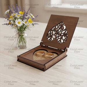 May include: A wooden ring box with a heart-shaped cutout design, holding two gold rings. The box is open, revealing a burlap-like interior. A small vase of flowers sits to the left.