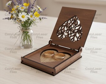 May include: A wooden ring box with a heart-shaped cutout design, holding two gold rings. The box is open, revealing a burlap-like interior. A small vase of flowers sits to the left.
