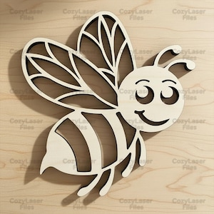 May include: A light-colored wooden cutout of a bee with a smiling face, two antennae, and large wings featuring a cut-out vein pattern. The bee has a rounded body with three horizontal stripes and is positioned against a light wood grain background.