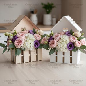 Puede incluir: Dos cajas de flores de madera, una natural y otra blanca, cada una con forma de casa con un corazón recortado. Las cajas están llenas de un vibrante arreglo de flores rosas, moradas y blancas, incluyendo rosas e hortensias. Las cajas están sobre una superficie de madera clara.