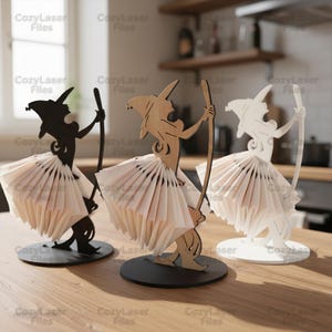 Puede incluir: Tres servilleteros con temática de brujas cortados con láser en negro, madera natural y blanco. Cada figura de bruja sostiene una escoba y una falda de servilletas dobladas. Los soportes están sobre bases redondas, ideales para Halloween.