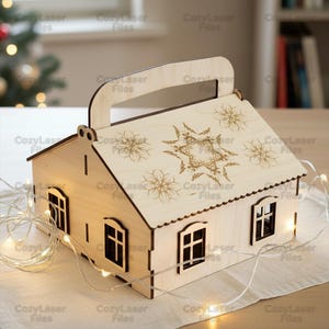 以下が含まれることがあります： 木製の家型のランタン。持ち手、窓、装飾的な屋根が付いています。屋根にはレーザーカットされた雪の結晶と星のデザインがあります。ランタンはストリングライトで照らされ、温かい光を放ちます。「CozyLaser Files」の文字が見えます。