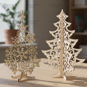 Op de afbeelding: Twee houten kerstboomversieringen. Eén boom heeft sneeuwvlokuitsparingen en een ster. De andere heeft een abstract ontwerp met uitgesneden vormen. Beide zijn gemaakt van lichtgekleurd hout en staan op een tafel.