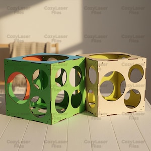 May include: Two wooden cube-shaped boxes with circular cutouts. One is painted green with blue and orange accents, the other is natural wood with yellow accents. The text "CozyLaser Files" is printed on the boxes.
