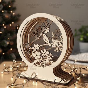 May include: A wooden, layered, circular decorative light featuring a bird perched on a flowering branch. The design is laser-cut with intricate details. The base is a curved shape, and the light is surrounded by a string of small white lights.