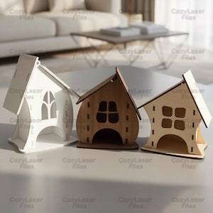Op de afbeelding: Drie houten huisvormige decoraties in wit, bruin en lichtbruin. Elk huis heeft een uniek raamontwerp en een kleine opening aan de basis. De tekst "CozyLaser Files" wordt herhaald op de afbeelding.