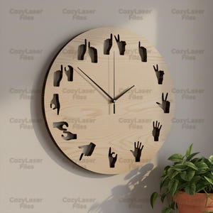 Puede incluir: Un reloj de pared redondo de madera clara con manecillas negras y gestos de manos recortados que representan números. El reloj tiene el texto "CozyLaser Files" repetido alrededor del perímetro.