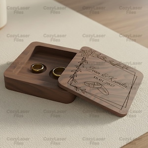 May include: A square dark brown wooden ring box with a sliding lid. The lid features a laser-engraved floral border, two interlocking rings, and the text "Ivan & Kamila 18.01.2027". The interior contains two circular indentations for holding rings.