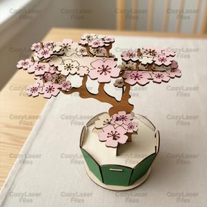 Può includere: Un bonsai in legno tagliato al laser con fiori di ciliegio rosa e bianchi. L'albero è in un vaso verde e bianco. Il design è intricato e dettagliato, perfetto per l'arredamento della casa.