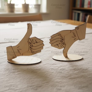 May include: Two wooden hand sculptures, one with a thumbs-up gesture and the other with a thumbs-down gesture. Both are laser-cut from light-colored wood and mounted on circular bases. The hands are detailed with etched lines.