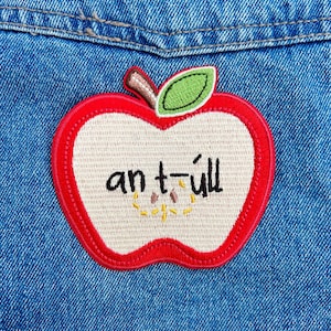 May include: Embroidered apple patch with a red border, a beige interior, and a green leaf. The patch features the text "an t-ull" in black. The patch is sewn onto a blue denim fabric.