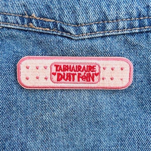 May include: A pink embroidered patch in the shape of a bandage, with the text "TABHAIRAIRE DUIT FEIN" in red. The patch is on a blue denim background. The patch has small heart and dot details.