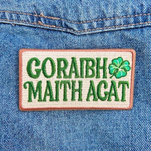 May include: Embroidered patch with the text "GORAIBH MAITH AGAT" in green, featuring a green shamrock on a cream background. The patch has a brown border and is set against a blue denim background.