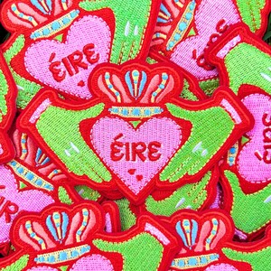 May include: A close-up of several embroidered felt patches in the shape of a Claddagh ring. The patches are red, green, and pink with the word "&Eacute;IRE" in the centre of the heart.