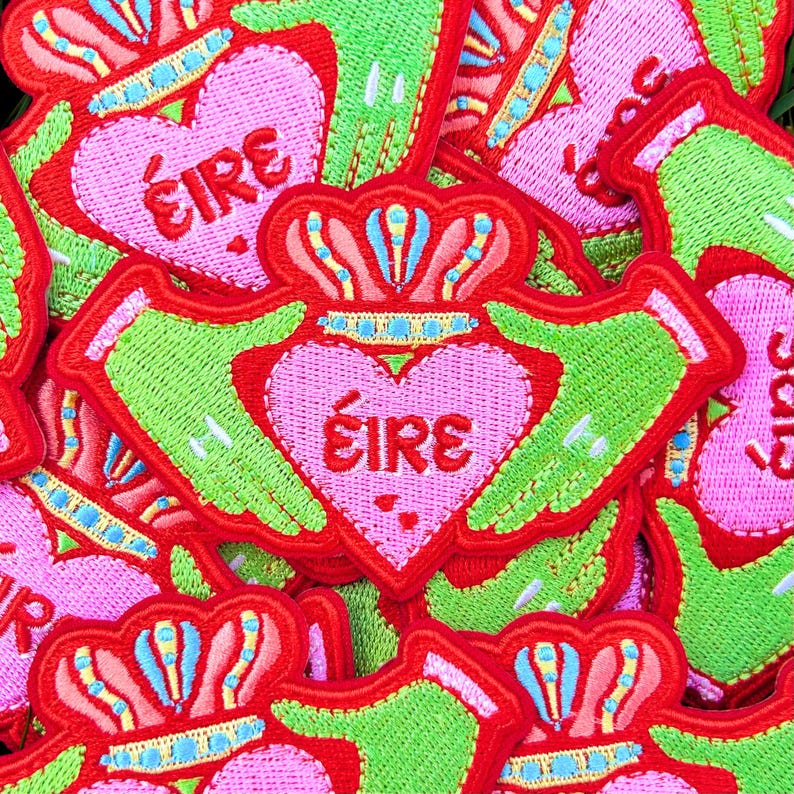 May include: A close-up of several embroidered felt patches in the shape of a Claddagh ring. The patches are red, green, and pink with the word "&Eacute;IRE" in the centre of the heart.
