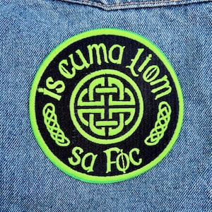 May include: A round, black patch with neon green trim and lettering. The patch features the text "is cuma liom sa Foc" in a circular pattern around a Celtic knot design. The patch is affixed to a denim jacket.