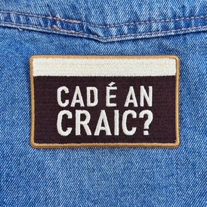Könnte beinhalten: Ein rechteckiger Aufnäher mit dem Text "CAD É AN CRAIC?" in weißen Buchstaben auf dunklem braunen Hintergrund. Der Aufnäher hat einen beigen Rand und einen weißen oberen Abschnitt, vor blauem Denim-Stoff.