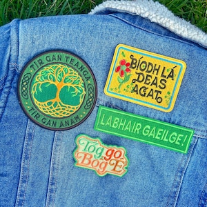 May include: A blue denim jacket with a white sherpa collar is adorned with four embroidered patches. The patches feature text in green and orange, and include a tree of life design and floral elements.