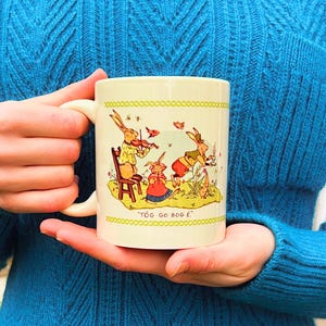 May include: A white ceramic mug with a handle features a whimsical illustration of three rabbits playing music. The rabbits are dressed in colorful clothing and are surrounded by flowers and birds. The text "TÖG GO BOG É" is printed on the bottom of the mug.