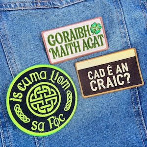 May include: Three embroidered patches with Gaelic text are affixed to a denim jacket. The patches feature green and white text, a shamrock, and Celtic knot designs. The patches are in various shapes, including rectangular and circular.